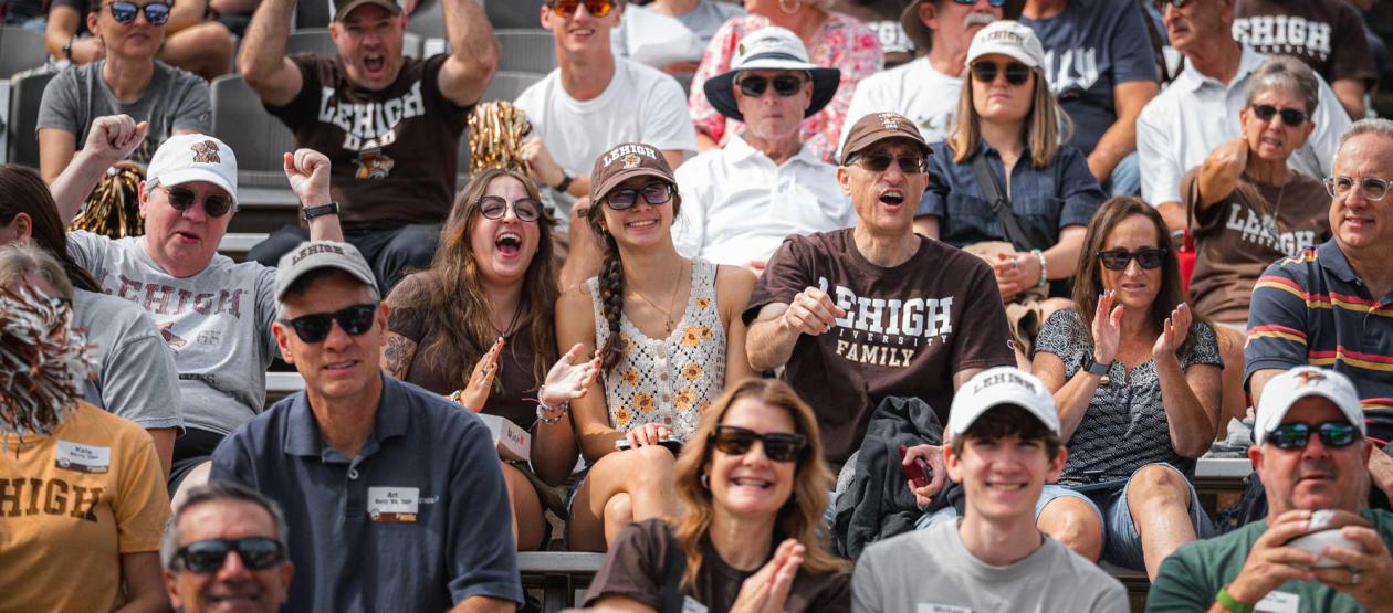 A crowded stadium bleacher filled with enthusiastic fans wearing brown and white Lehigh University gear cheering during a football game.