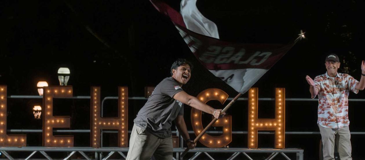 A Lehigh University student triumphantly waves a maroon and white class flag on a stage at night, with a large, lit-up "LEHIGH" sign in the background. An alumnus stands to the side, clapping enthusiastically.