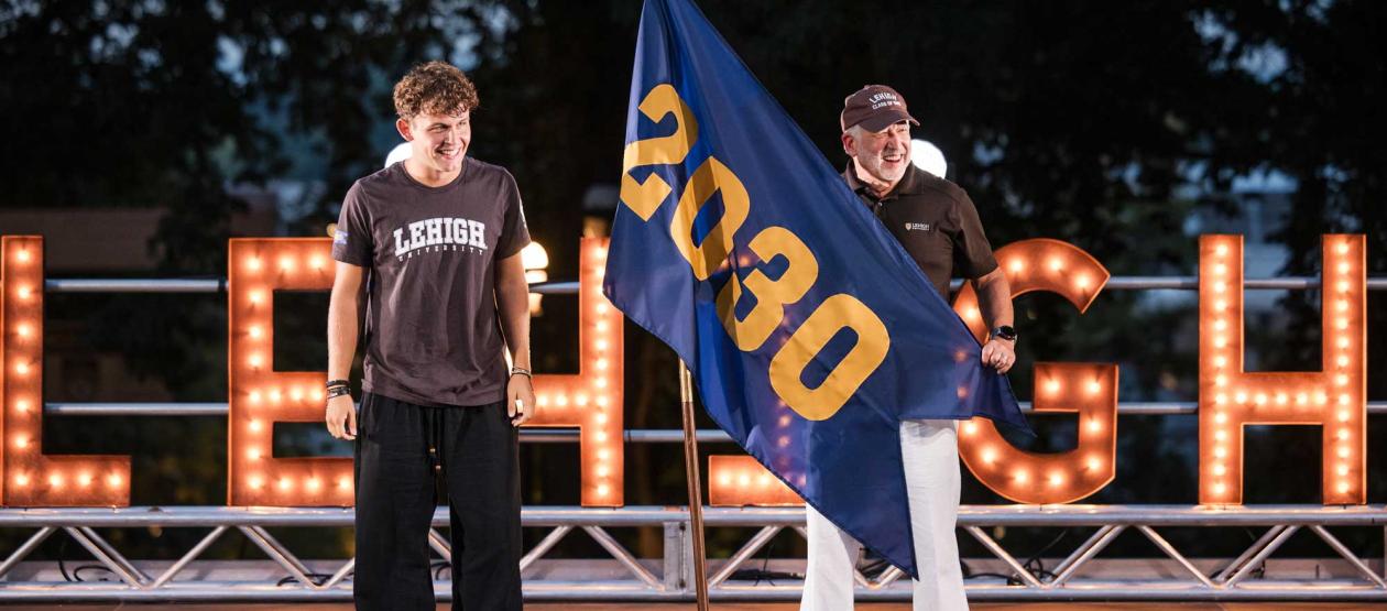 Two men stand on a stage in front of large, illuminated "LEHIGH" marquee letters holding a blue flag that reads "2030."
