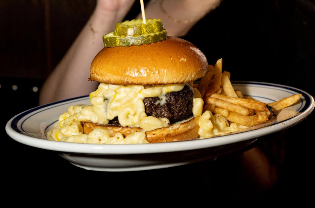 A mac and cheese burger resting on a plate surrounded by crunchy french fries