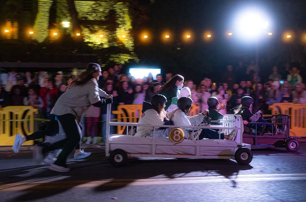 One team of Lehigh students race their bed-shaped cars down Packer Avenue with a crowd of spectators cheering in the background.