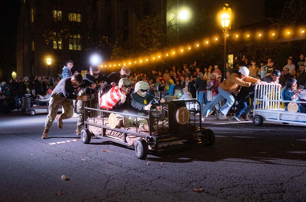 Two groups of Lehigh students race their bed-shaped cars down Packer Avenue with a crowd of spectators cheering in the background.