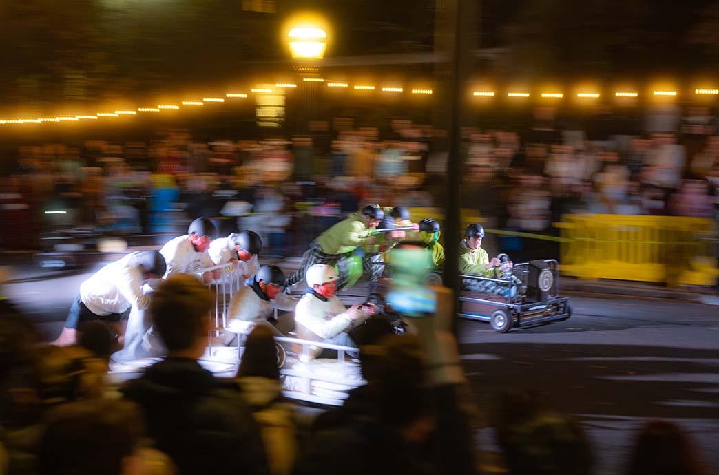The blur of motion is visible as two bed-shaped racing cars are pushed down the street by students.