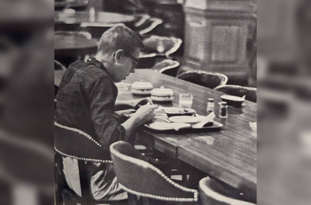 Black and white photo shows a student sitting at a dining hall table hunched over a plate of food.