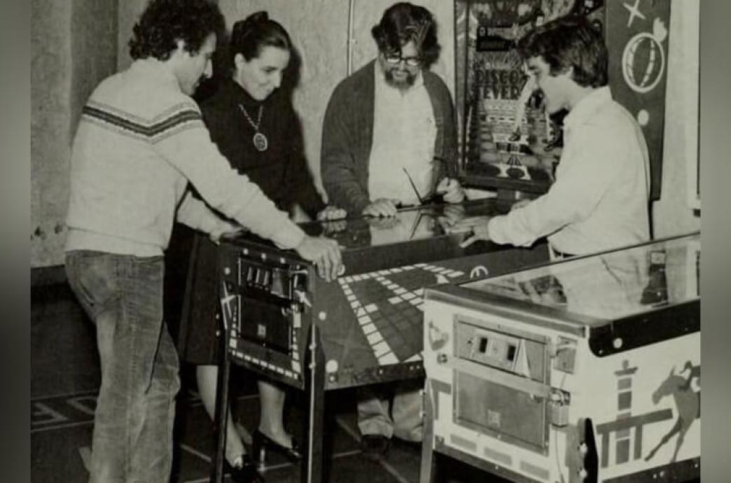 Black and white photo shows four students in 1980 gathered around a pinball machine watching one play the game.