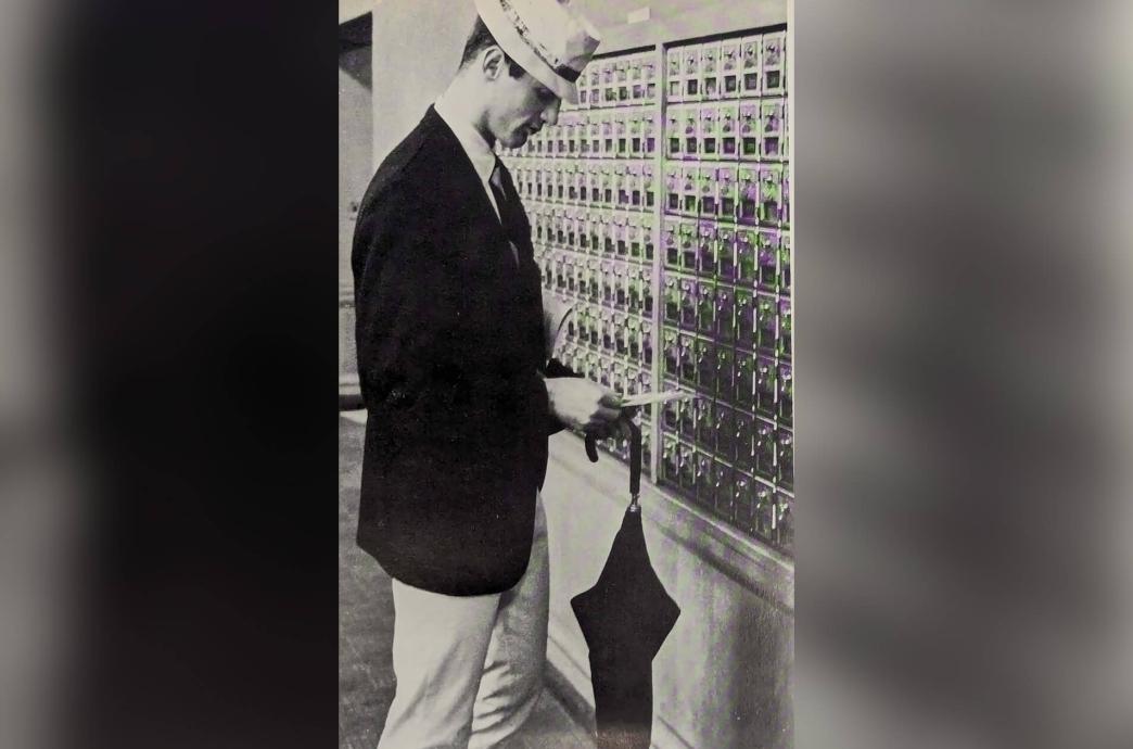 Black and white photo from the 1963 yearbook shows a man wearing a hat and blazer standing at a wall of mailboxes looking down at his mail.