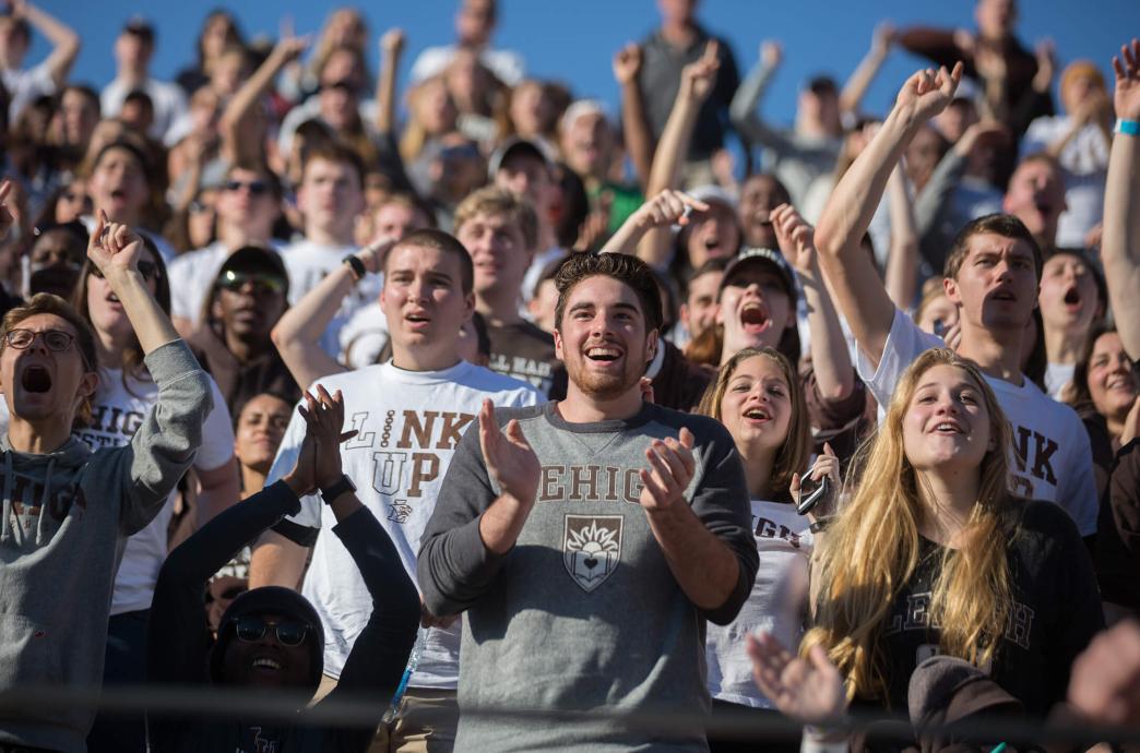 Fans stand and applaud in the stands at a football game wearing Lehigh attire.