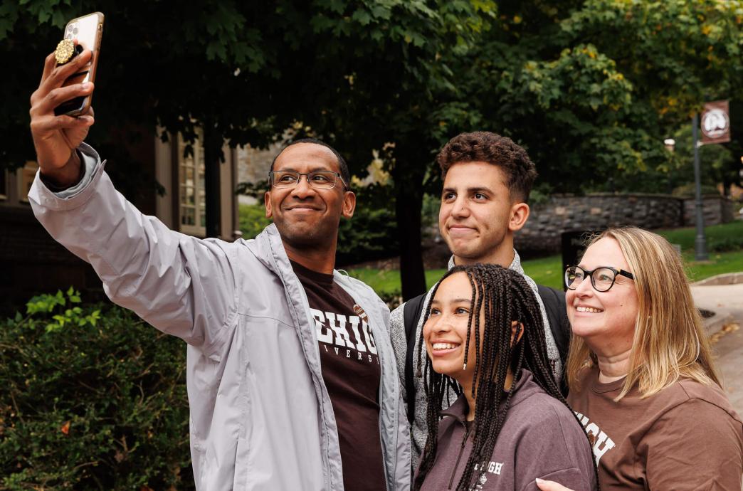 Family of four dressed in Lehigh gear and smiling up at a cell phone as they stop to take a selfie on campus.