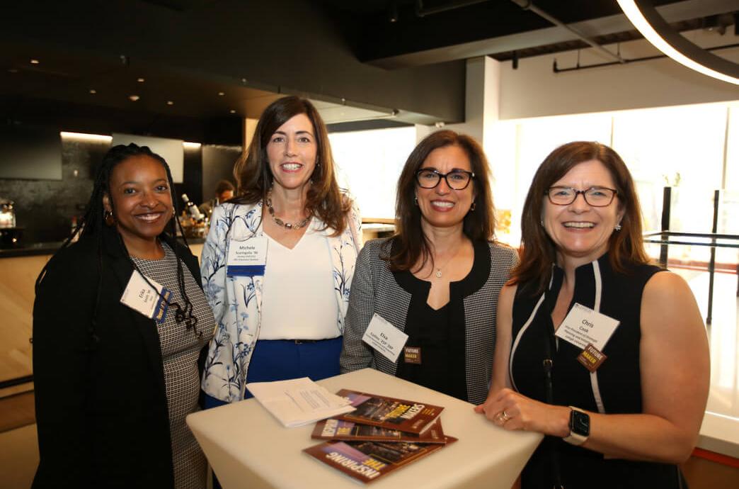 Four women with name tags stand at a high top cocktail table smiling at the camera