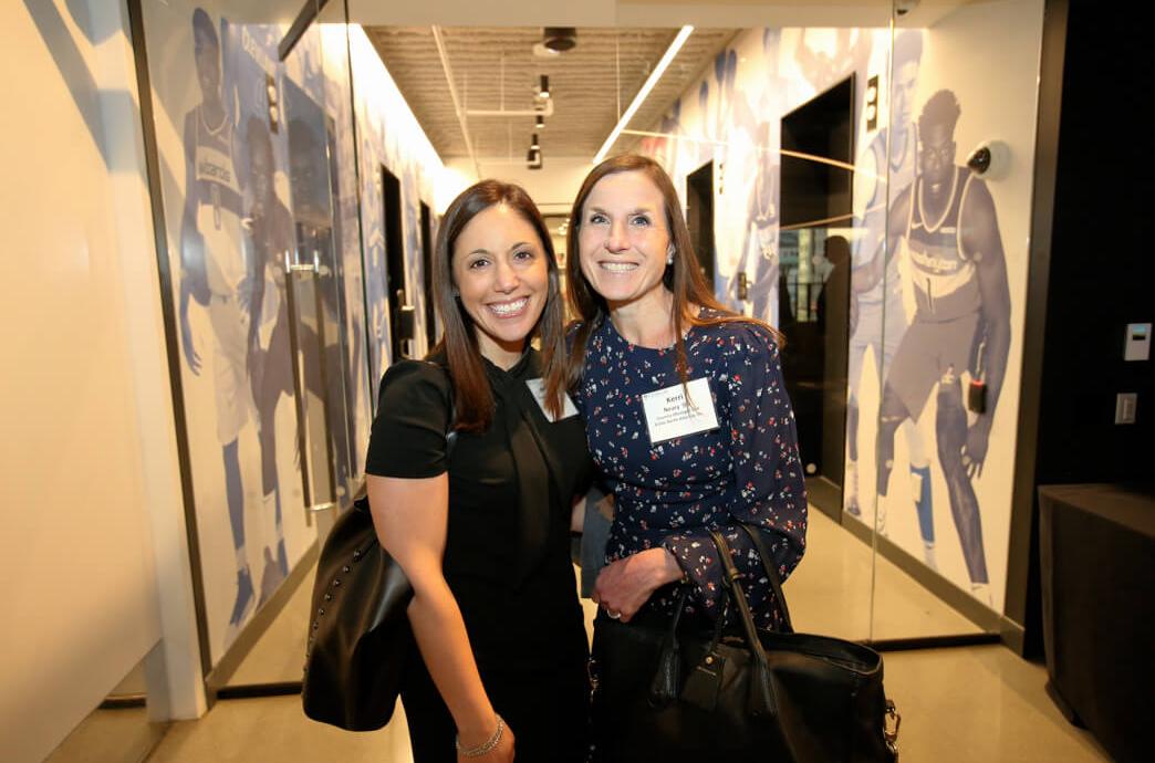 Two women smile for the camera while standing in a corridor lined with floor to ceiling photo murals of basketball players 