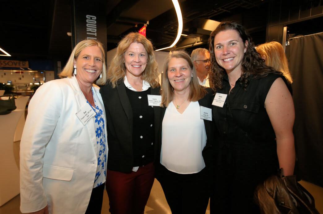 Four women wearing name tags stand shoulder-to-shoulder and smile at the camera at an event.