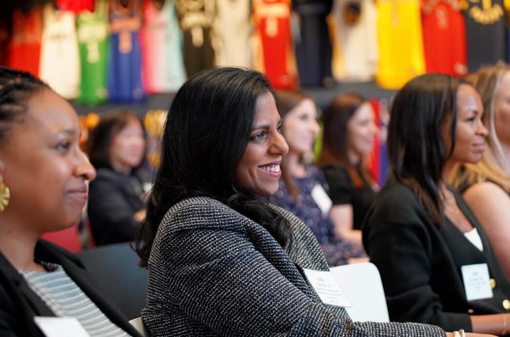Female audience sits facing one direction and the camera focuses on one women smiling with rows of basketball jerseys against the wall