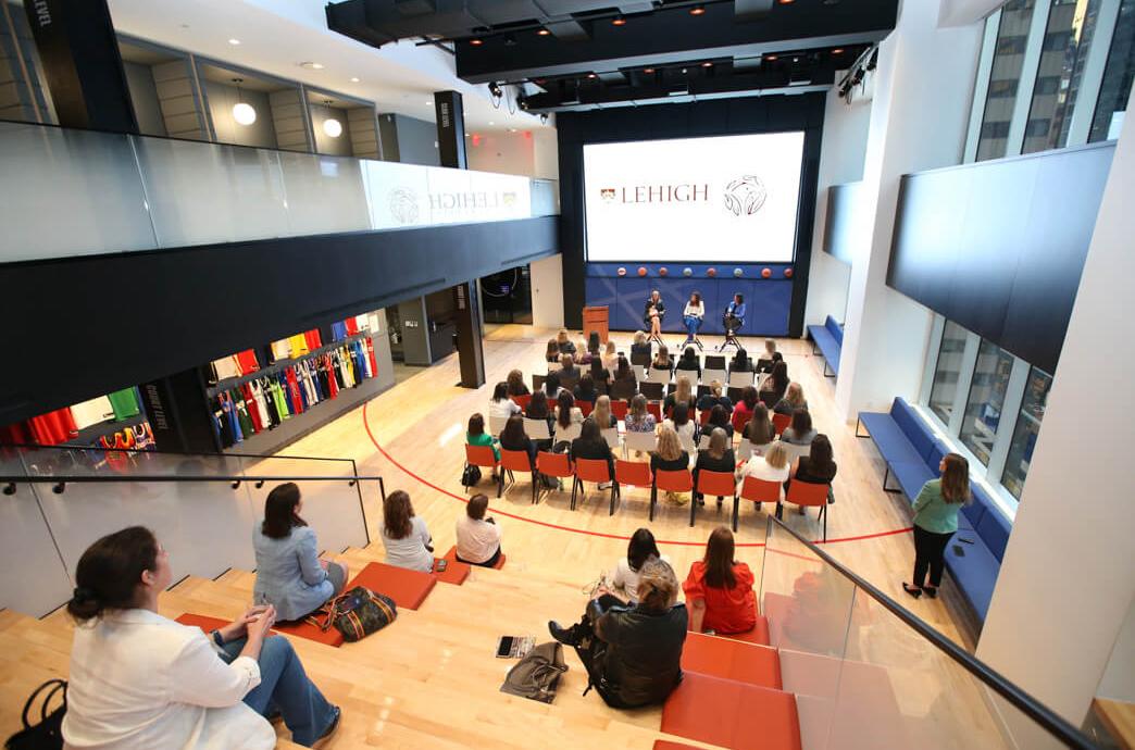 A room showing an audience from behind assembled in casual stadium seating and chairs at the front with a screen that says "Lehigh"