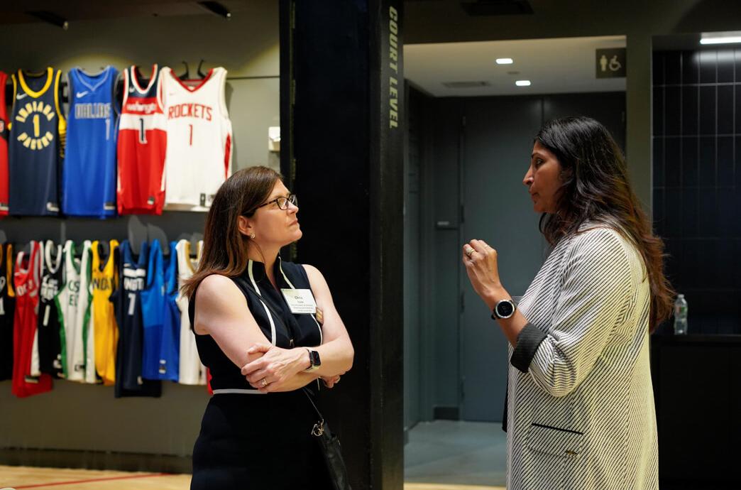 Two alumnae speak intently in a large room with a row of basketball jerseys int he background.