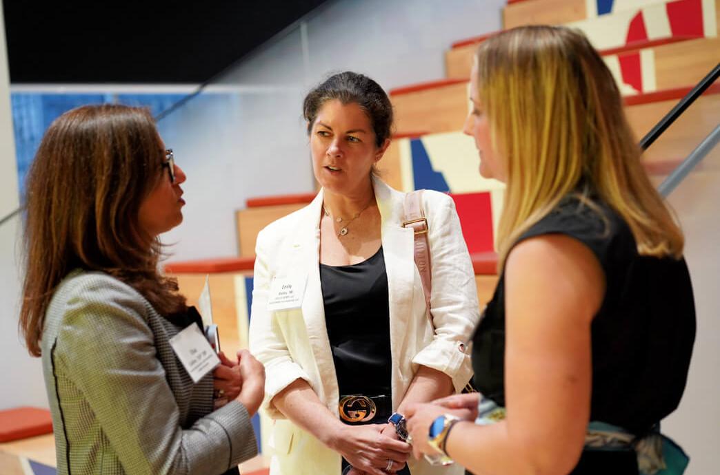 Three alumnae engage in conversation in a room that features casual stadium seating behind them.