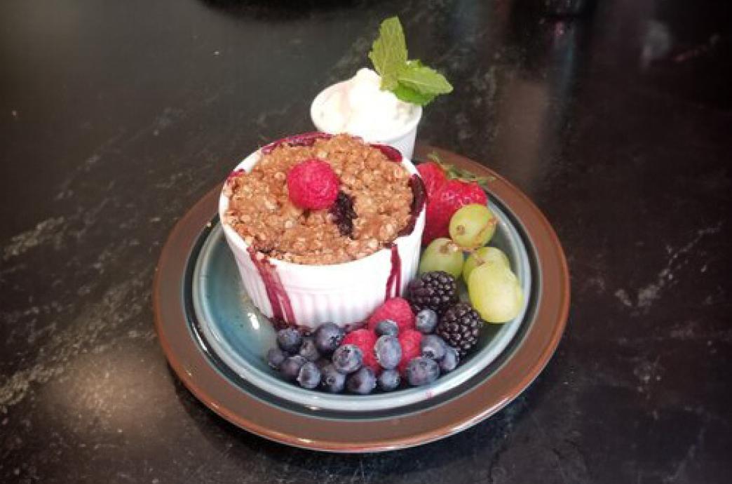 A crumble-style dessert in a ramekin, garnished with berries, whipped cream, and a sprig of mint.