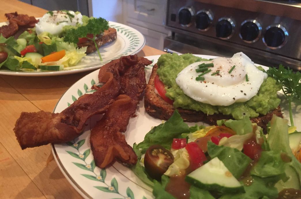 Two plates serving a green salad, two slices of bacon, and a piece of whole grain toast topped with avocado and a poached egg.