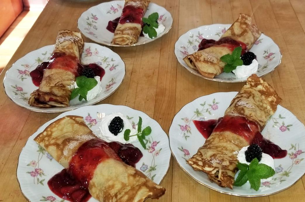 Five white china plates sit on a table, each staged with one crepe, drizzled with red sauce, whipped cream, and a sprig of mint.
