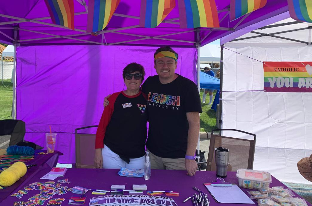 Two Pride Center staff stand at under purple tent with pride flags hanging above them.