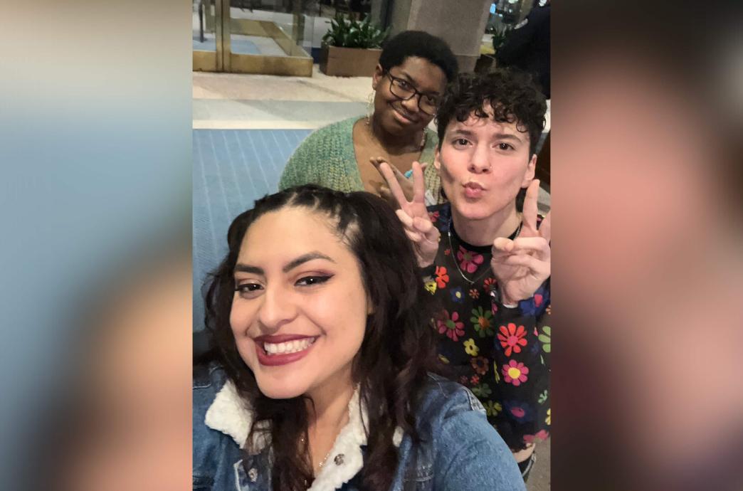 Three students pose for a selfie with one in the middle making a kiss face and holding up peace signs with his hands.