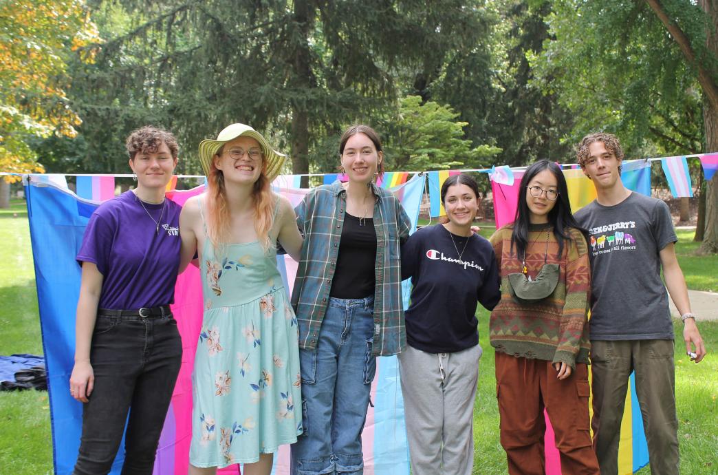 Six students stand at a Pride event on campus with Pride flags hung behind them.