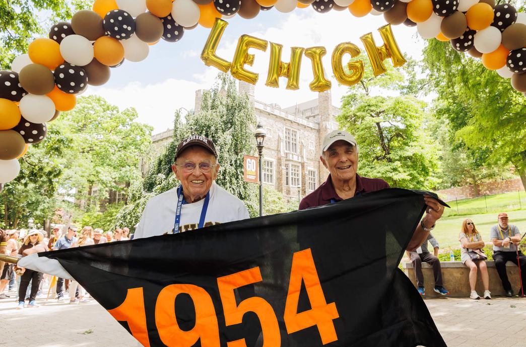 Two alumni from the class of 1954 hold their black flag with orange numbers as they pose under a balloon arch.