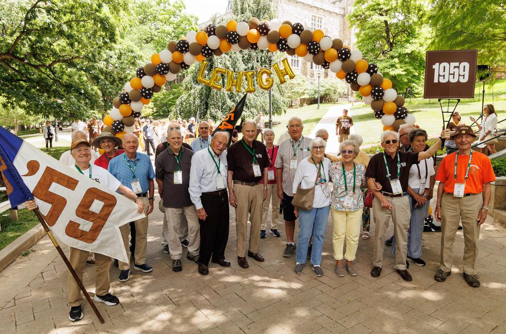 Alumni from 1959 pose under a balloon arch holding flags and signs with their class year.
