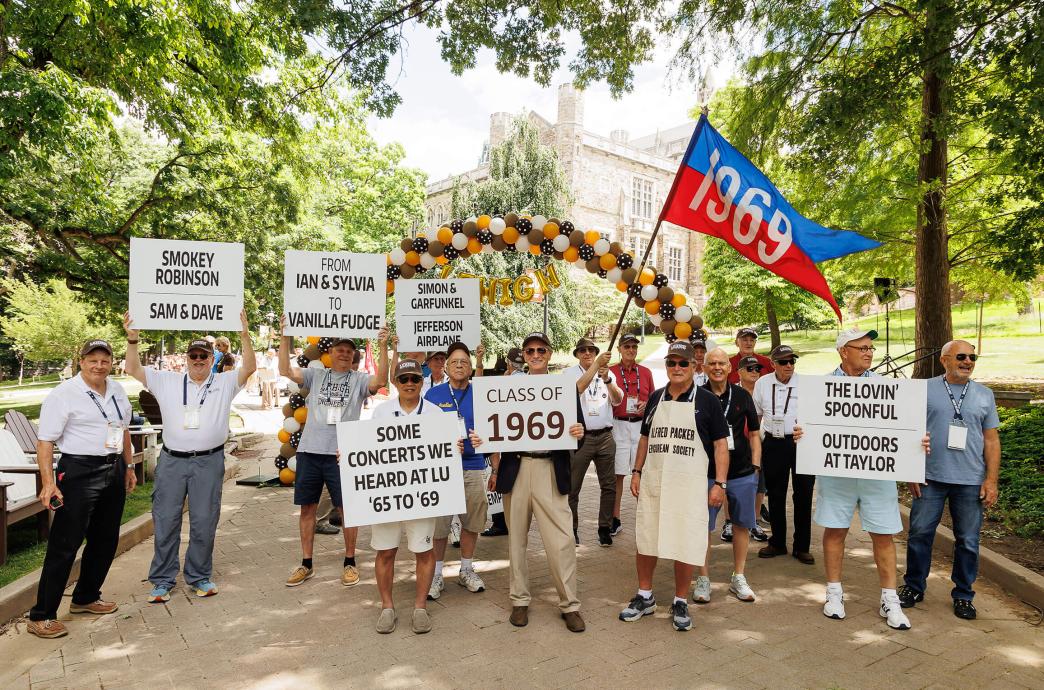 The Class of 1969 hold their red and blue class flag and signs that read the names of concerts they attended as students.