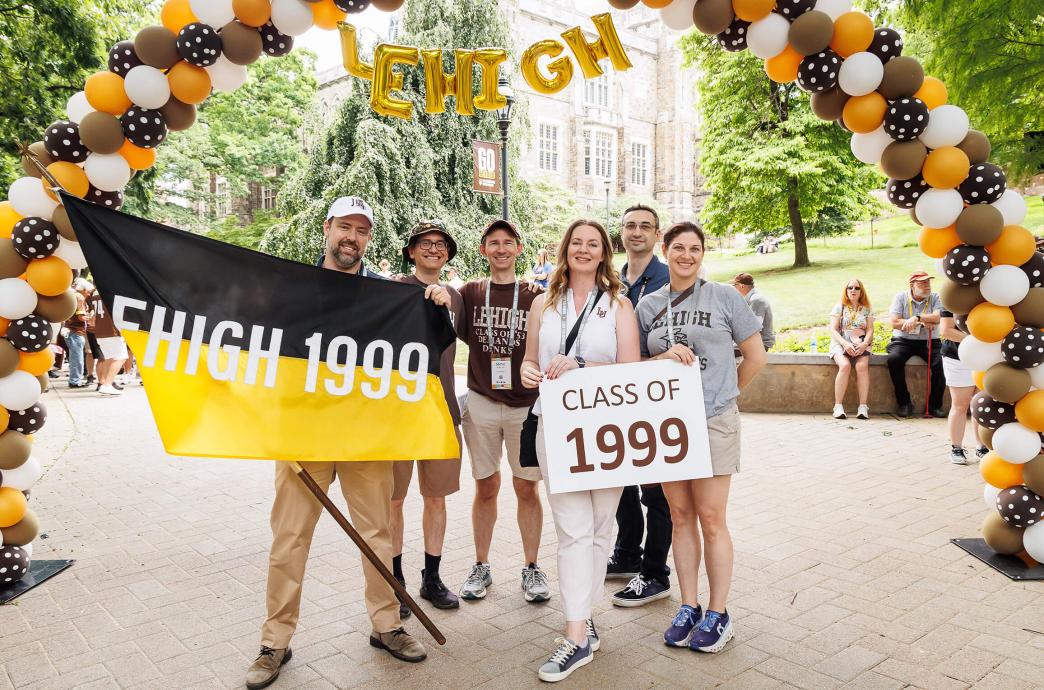 Six alumni from the class of 1999 pose under a balloon arch holding their black and yellow class flag and a sign with their class year.