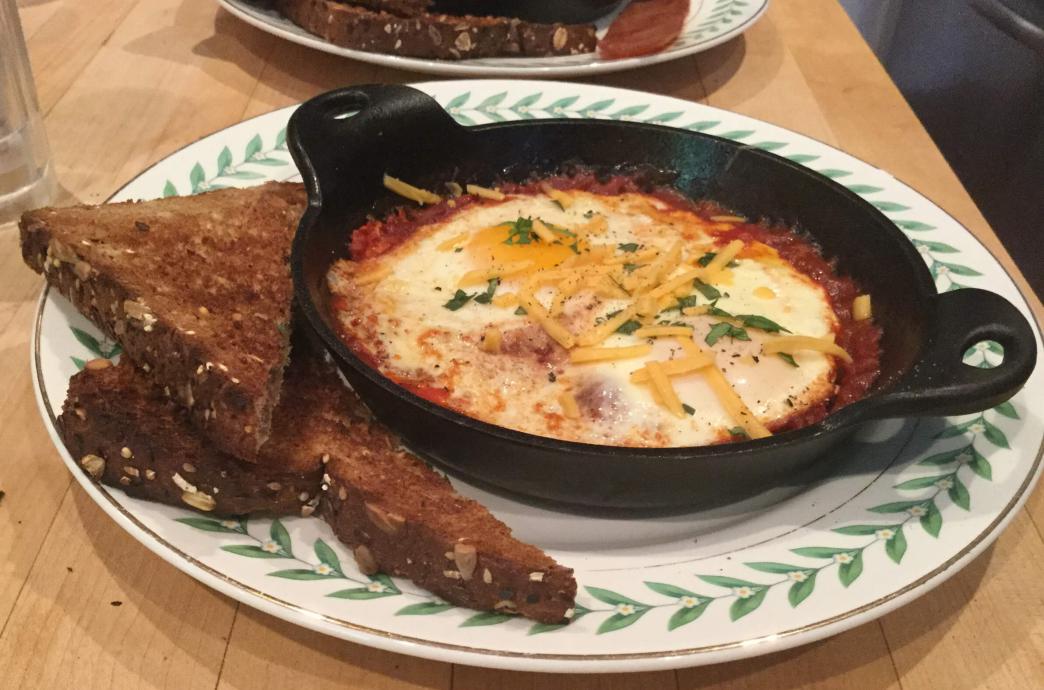 A cast iron pan, filled with eggs poached in red sauce and sprinkled with cheese and served with two slices of whole grain toast.