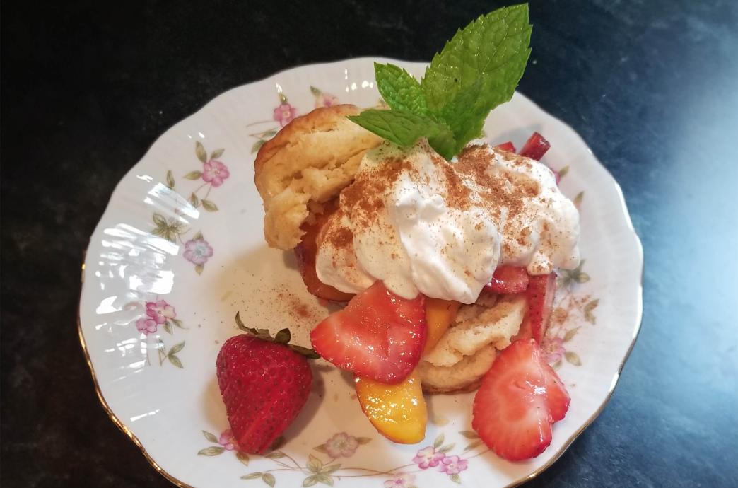 A shortcake with whipped cream, strawberries and peaches, a sprig of mint and cinnamon sits on a white china with floral details.