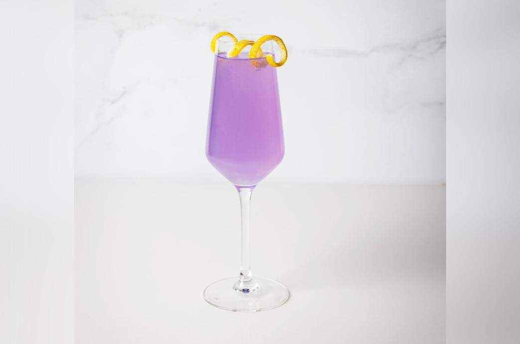 A lavender colored cocktail in a stemmed cocktail glass with tall straight sides, garnished with a citrus peel.