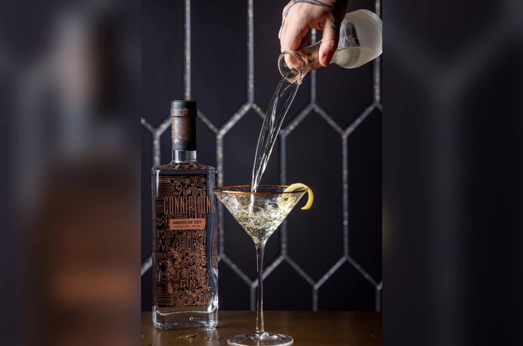 A gin bottle stands beside a gold-rimmed martini glass garnished with a peel while a clear cocktail is poured from a glass carafe.