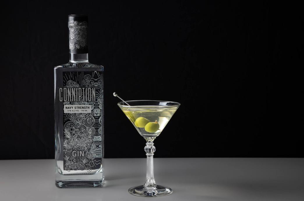 A bottle of gin stands beside a martini glass garnished with olives, shot against a dark background.