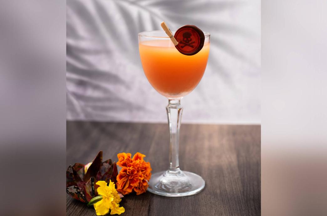 Flowers sit beside a coral colored cocktail in a stemmed cocktail glass garnished with a skull and crossbones medallion.