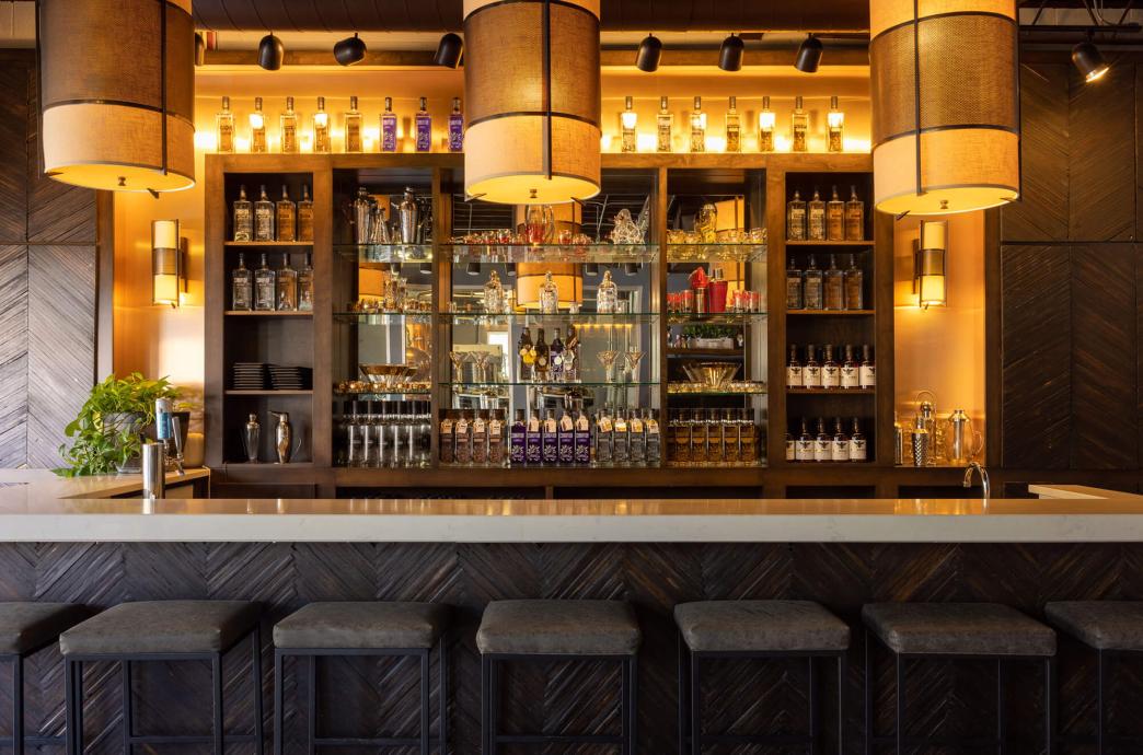 An intimate, warmly lit room with a white bar top and lit back bar featuring different bottles of liquor.