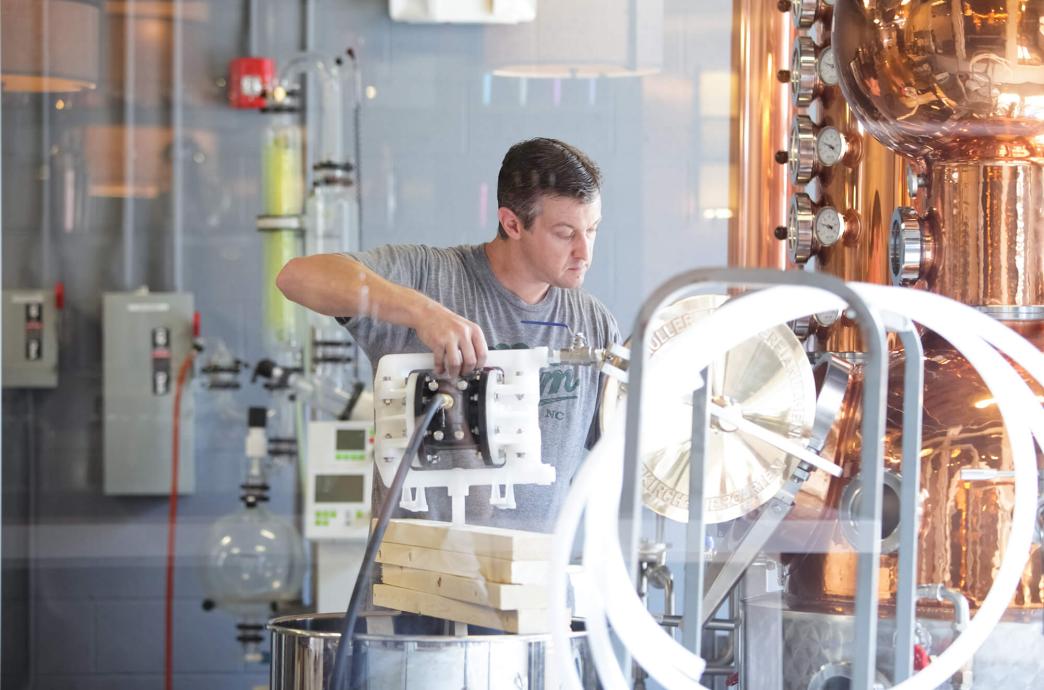 Man stands in a distillery, surrounded by equipment and barrels, working carefully.