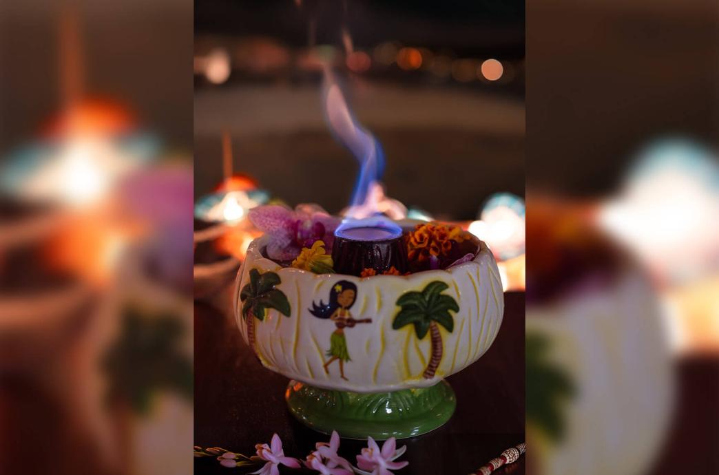 A flamboyant cocktail blazes with a blue flame, garnished with tropical flowers in a porcelain bowl with Hawaiian designs.