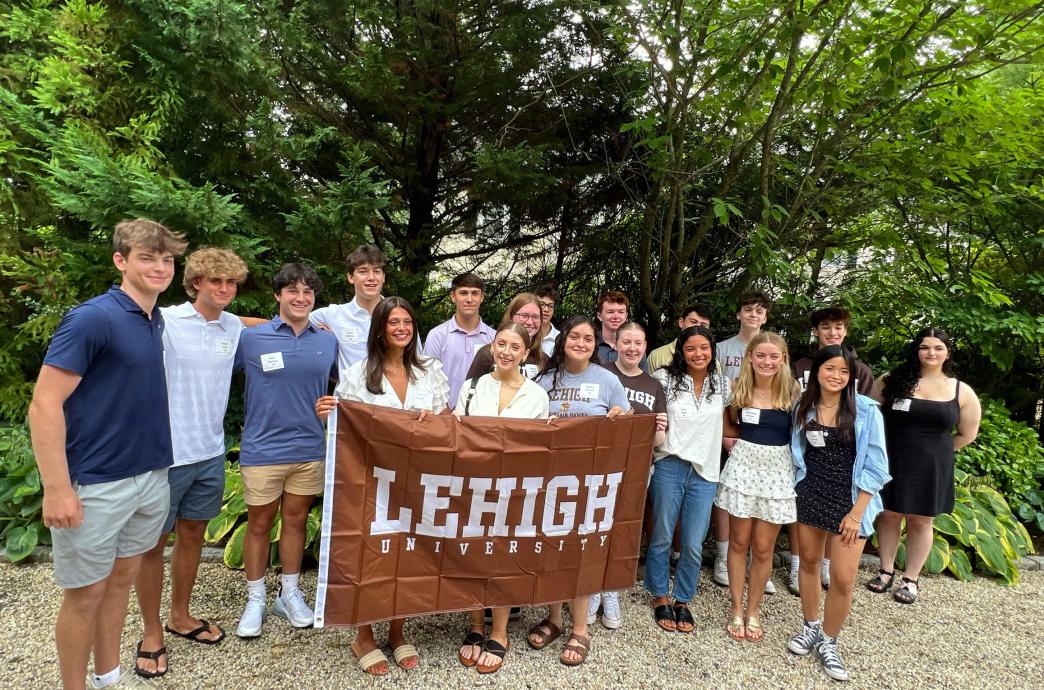 Students standing in front of green foliage while smiling and holding a brown Lehigh flag.