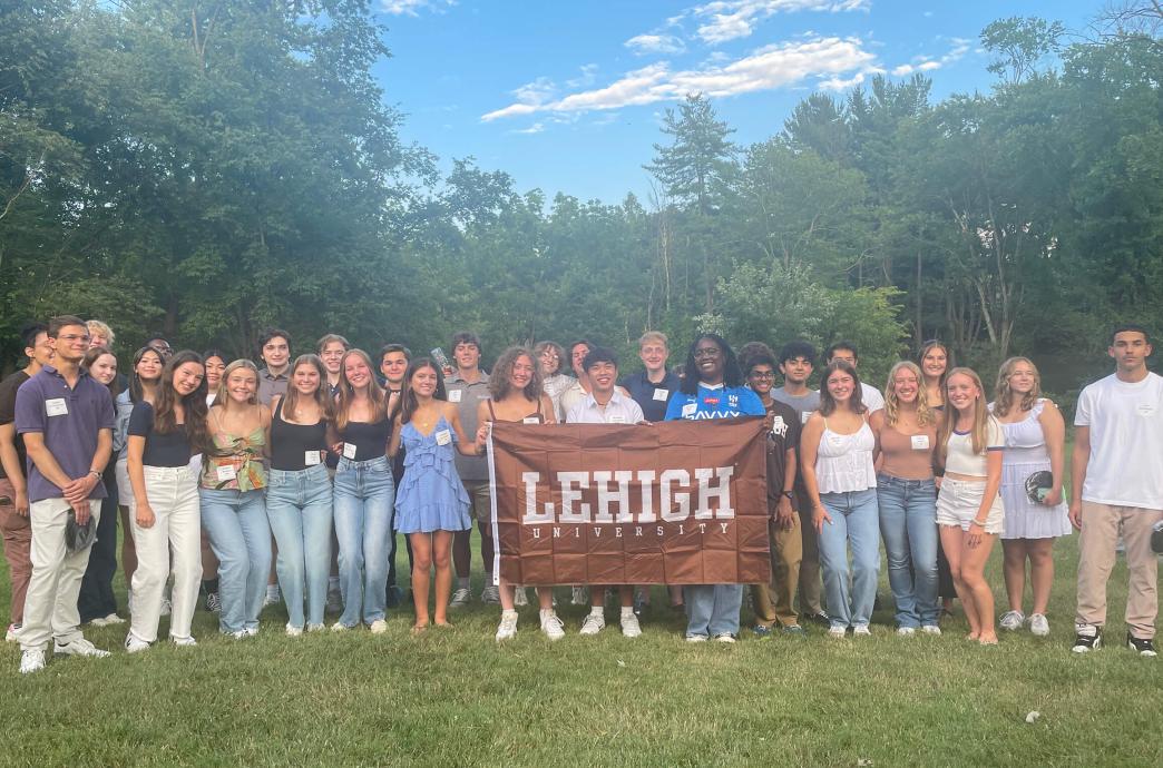 Students standing in front of green foliage while smiling and holding a brown Lehigh flag.