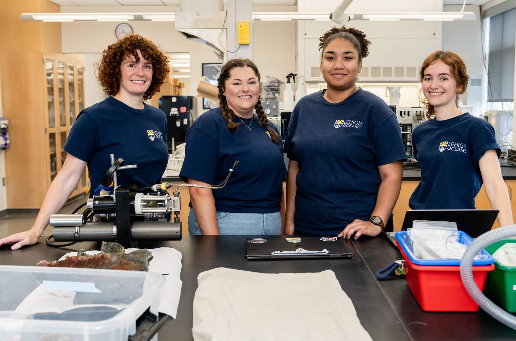 Four women stand in a desk in a lab, all wearing navy blue Lehigh Oceans shirts.