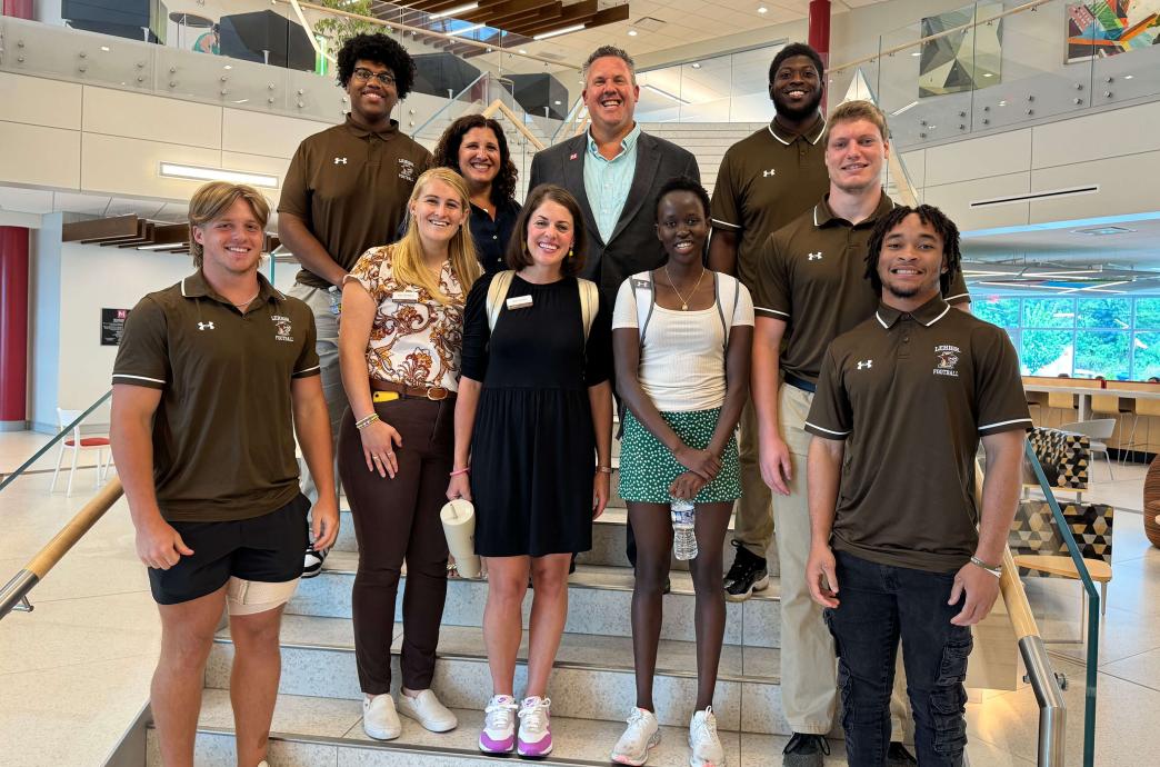 A group stands on a staircase inside a company lobby, some wearing brown athletics shirts.