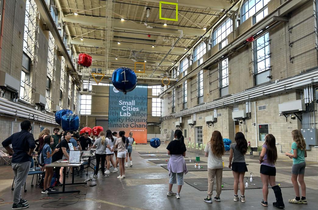 Middle school girls fly blimp-style drones through obstacles in a warehouse style building with high ceilings.