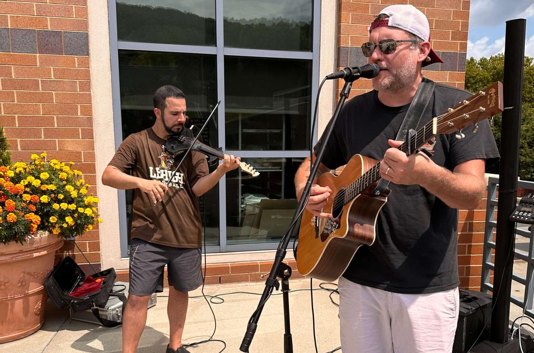 Both outdoors, one musician wears a brown Lehigh baseball shirt and plays a violin; another plays a guitar at a microphone.