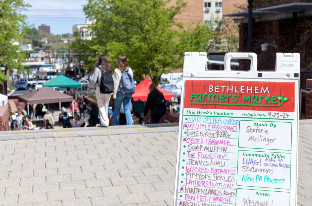 A standing sign on the ground lists the offerings of the farmer's market with the market going on in the background.
