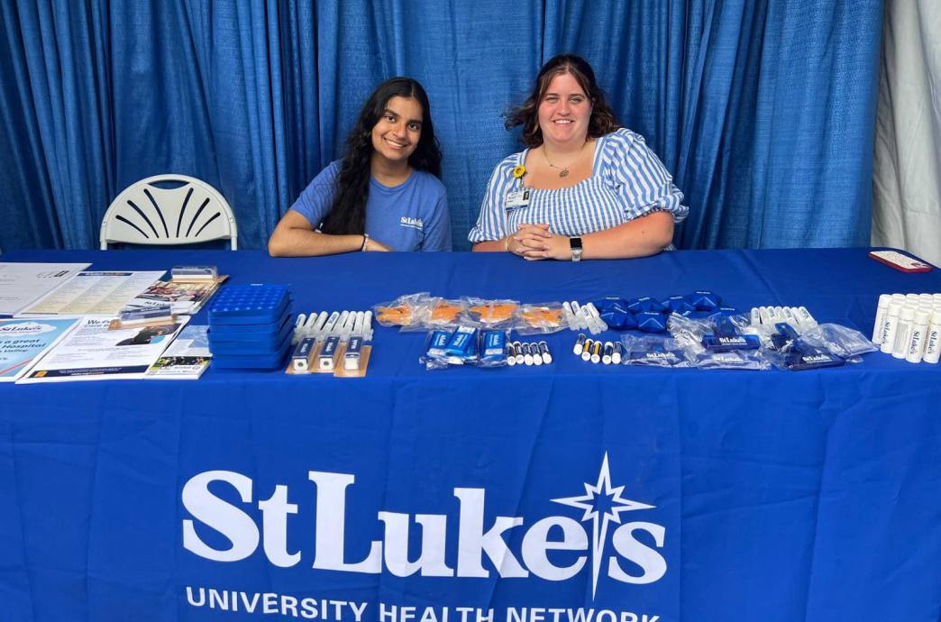 Two female interns sit at a table draped in blue signage and swag for St. Luke's University Health Network.