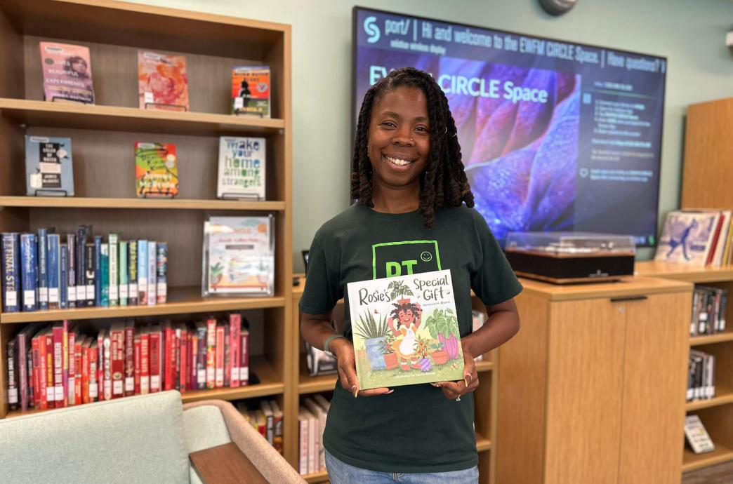 Woman poses in front of a bookcase and smartboard holding a children's book.
