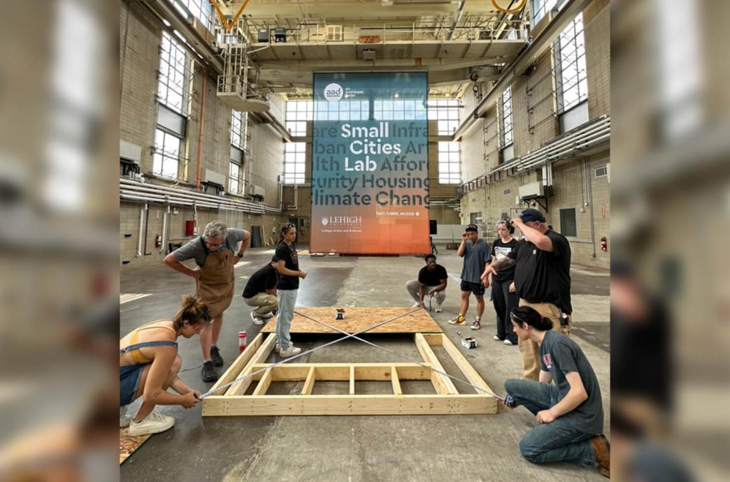 Students build a wooden structure on the ground in a warehouse-style room with a sign behind them that reads Small Cities Lab.