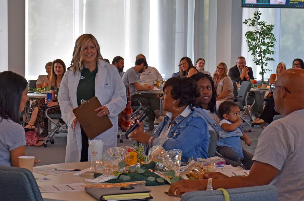 A woman wearing a lab coat stands in the center of a room filled with seated attendees.