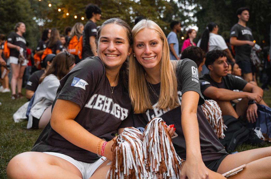 Two students wearing Lehigh shirts sit on the ground holding pompoms, surrounded by fellow students.